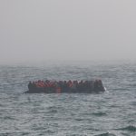 Crowded inflatable boat carrying many people in orange life jackets on rough sea.