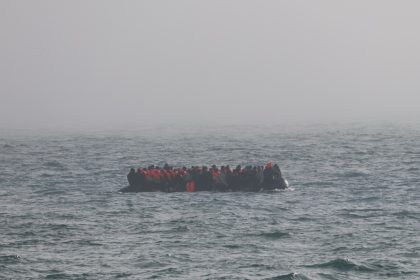 Crowded inflatable boat carrying many people in orange life jackets on rough sea.
