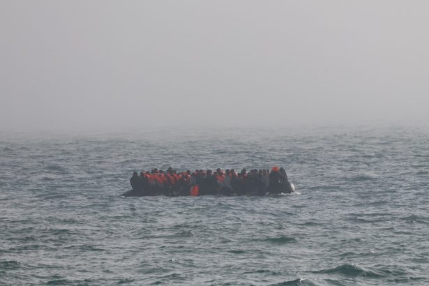 Crowded inflatable boat carrying many people in orange life jackets on rough sea.