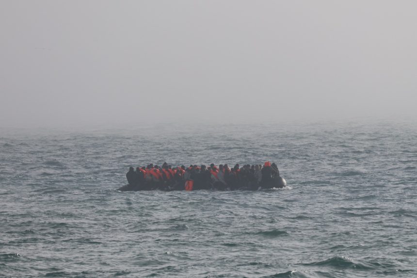 Crowded inflatable boat carrying many people in orange life jackets on rough sea.