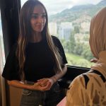 9a309c5f-a308-44f6-b20a-34d9f87799e8 - The Afghan Times Two women chatting in an elevator by a window, one in a black shirt and jeans, the other wearing a patterned hijab.